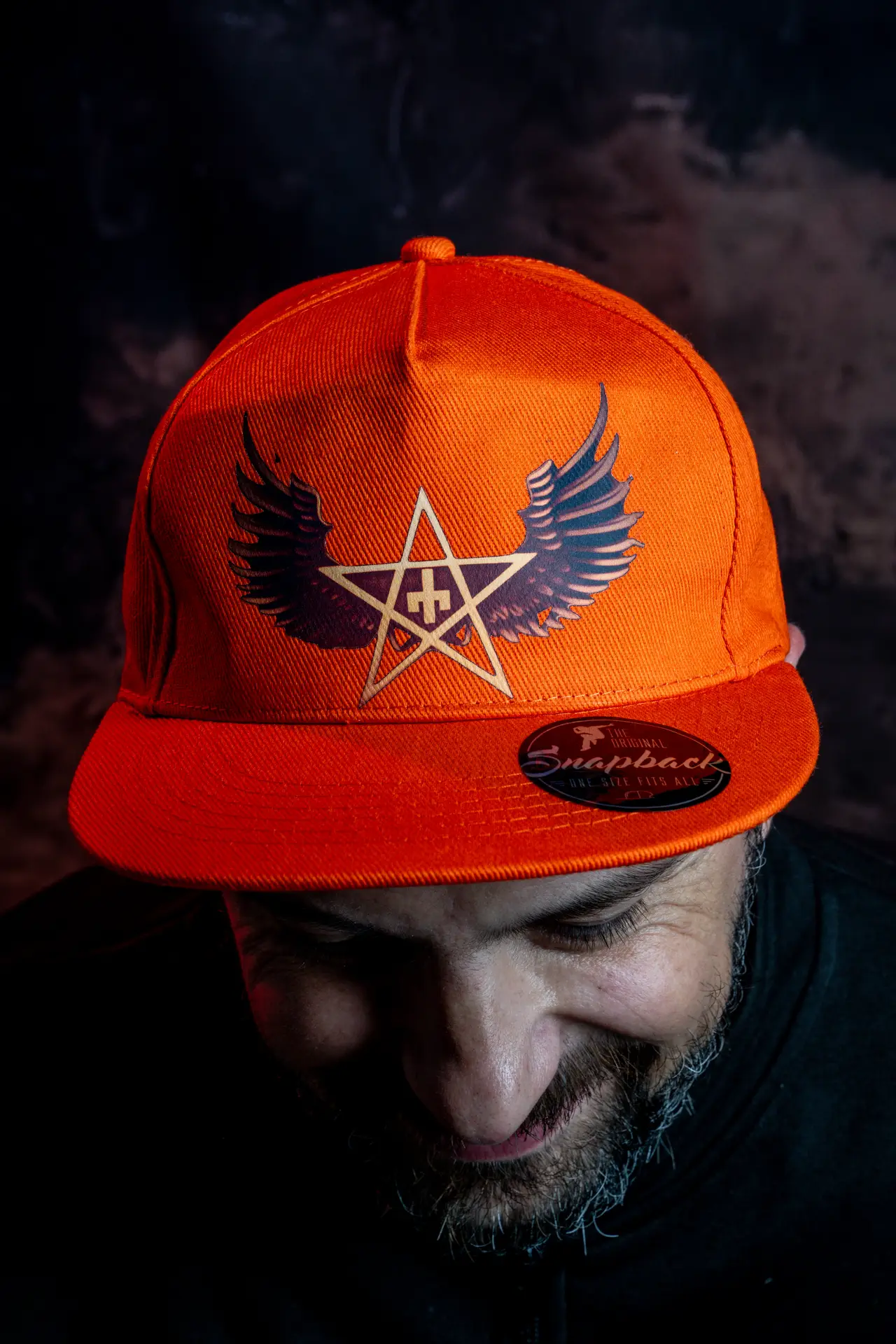 A close-up image of a man wearing an orange snapback cap featuring a design of wings and a star.